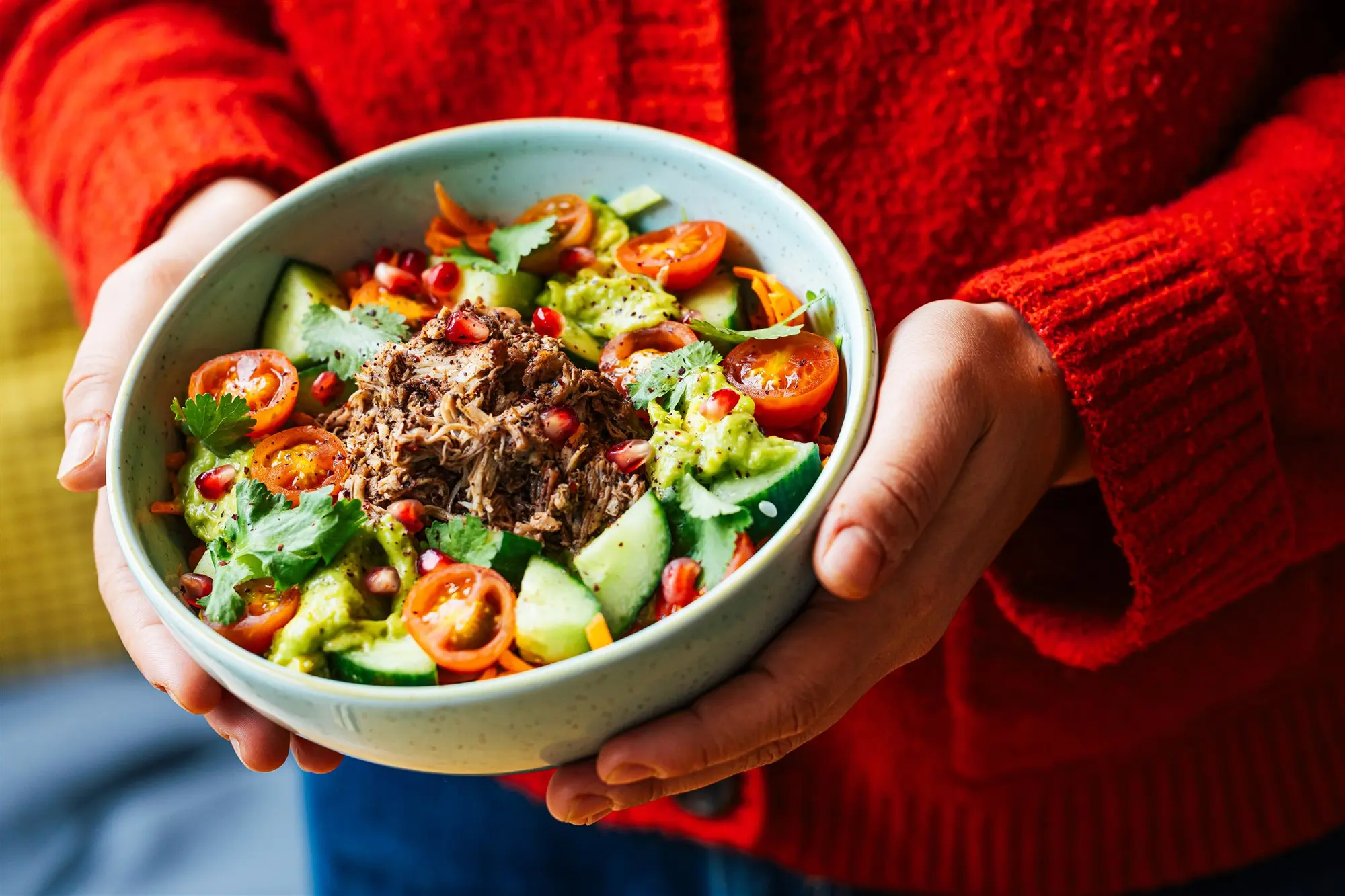 Hands in a red knit jumper holding a Peeld chicken shawarma bowl with tomato, cucumber, avocado and pomegranate